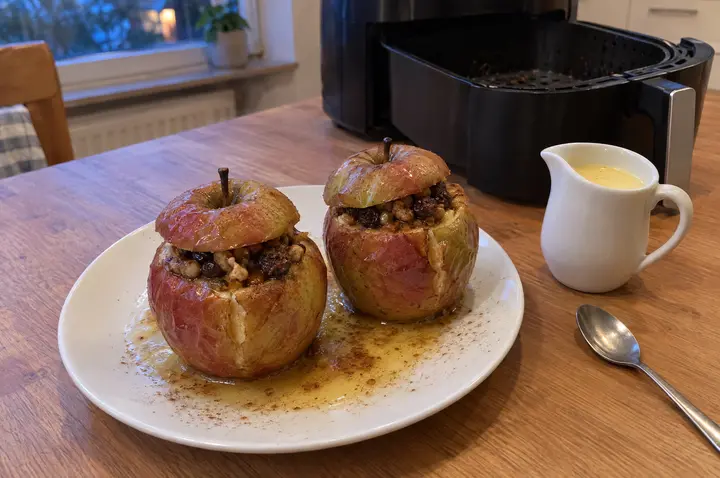 Zwei gefüllte Bratäpfel mit Walnüssen und Rosinen und aufgesetztem Deckel auf einem weißen Teller mit Zimt und Apfelsaft, daneben ein Kännchen Vanillesoße; im Hintergrund die geöffnete Heißluftfritteuse