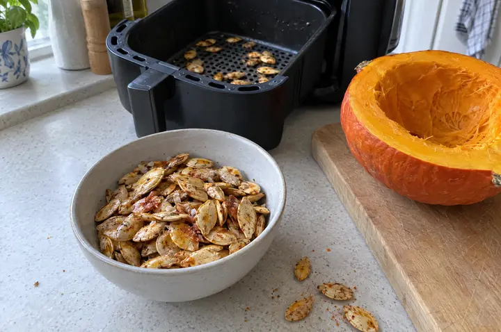 Geröstete Hokkaido-Kürbiskerne mit Schale, mit Salz und Paprika gewürzt, in einer hellen Keramikschüssel; daneben ein ausgehöhlter halber Hokkaido-Kürbis, dahinter der Korb der Heißluftfritteuse mit weiteren Kernen