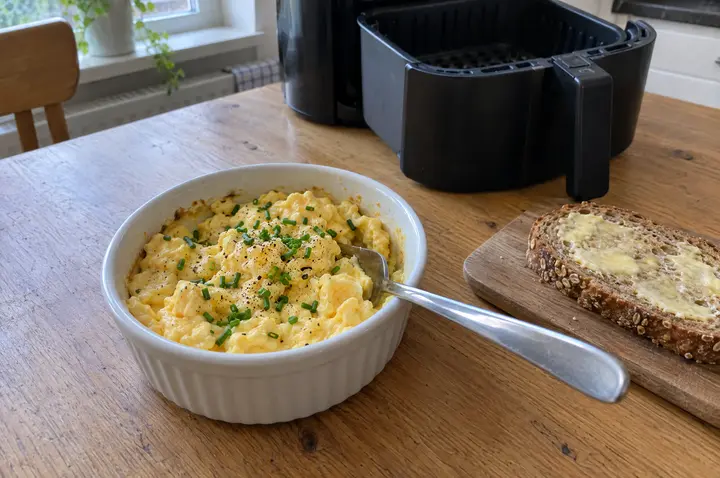 Cremiges, lockeres Rührei mit Schnittlauch und schwarzem Pfeffer in einer kleinen weißen Auflaufform mit Gabel, daneben eine Scheibe Vollkornbrot mit Butter; im Hintergrund die geöffnete Heißluftfritteuse